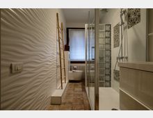 Bagno moderno e stretto con parete in piastrelle bianche ondulate, pavimento in gres e box doccia in vetro affiancato da una parete di mattoni di vetro. In fondo si vedono WC sospeso e finestra con tenda, mentre a destra ci sono lavabo con mobile e rivestimenti a mosaico.