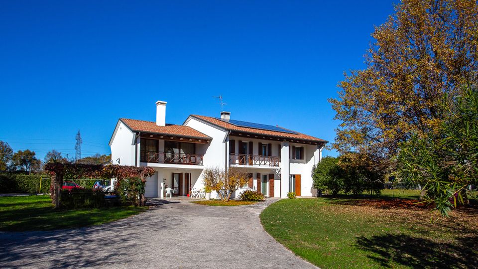 Casa a due piani intonacata di bianco con tetto in coppi rossi e balconi in legno, situata in un ampio giardino. Un vialetto asfaltato conduce all’ingresso, con alberi e vegetazione ai lati sotto un cielo sereno.