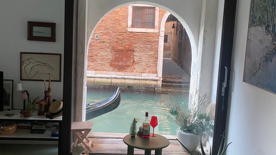 Interno di uno spazio moderno con pavimento lucido, decorato con una poltrona arancione, un tavolino e piante, attraverso una grande finestra ad arco che dà su un canale veneziano con una gondola visibile sull'acqua. Il soffitto presenta travi in legno bianche.