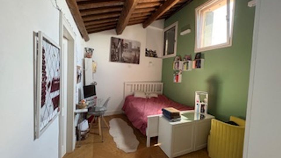 Camera da letto con soffitto in legno a travi a vista, parete verde e pavimento in parquet. Sono visibili un letto con copriletto rosso, una scrivania con sedia, mensole con libri e una finestra che illumina la stanza.