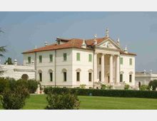 Villa storica in stile neoclassico con facciata bianca, portico con alte colonne e frontone decorato da statue, tetto in coppi e finestre con persiane verdi. Davanti si estende un ampio prato con siepi e giardino curato.