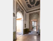 Interno di un edificio storico con pavimento in pietra, pareti con intonaci decorativi e due grandi aperture ad arco che conducono a un cortile luminoso e a una stanza con finestra. Soffitto a cassettoni dipinto con motivi geometrici e cornici ornate sopra i portali.