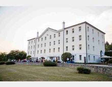 Grande edificio bianco a più piani con tetto a falde e molte finestre rettangolari, affacciato su un ampio prato e un muretto in pietra. Davanti all’ingresso si vedono gazebo e aree all’aperto con vegetazione e alberi sullo sfondo.