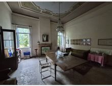 Interno di un salone d’epoca con soffitto alto decorato a stucchi e lampadario in cristallo, pavimento in marmo e grandi finestre con tende bianche. Al centro c’è un tavolo in legno, lungo una parete un divano e una fila di cornici, e in fondo un camino con specchio.