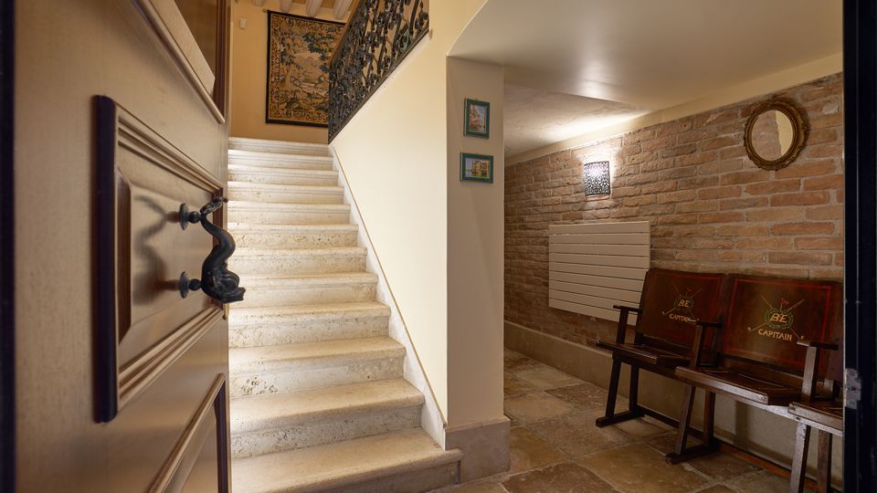 Ingresso interno con scala in pietra chiara e corrimano in ferro battuto che sale al piano superiore, visto da una porta socchiusa. A destra, piccolo corridoio con parete in mattoni a vista, panchina in legno, applique a parete e specchio rotondo.