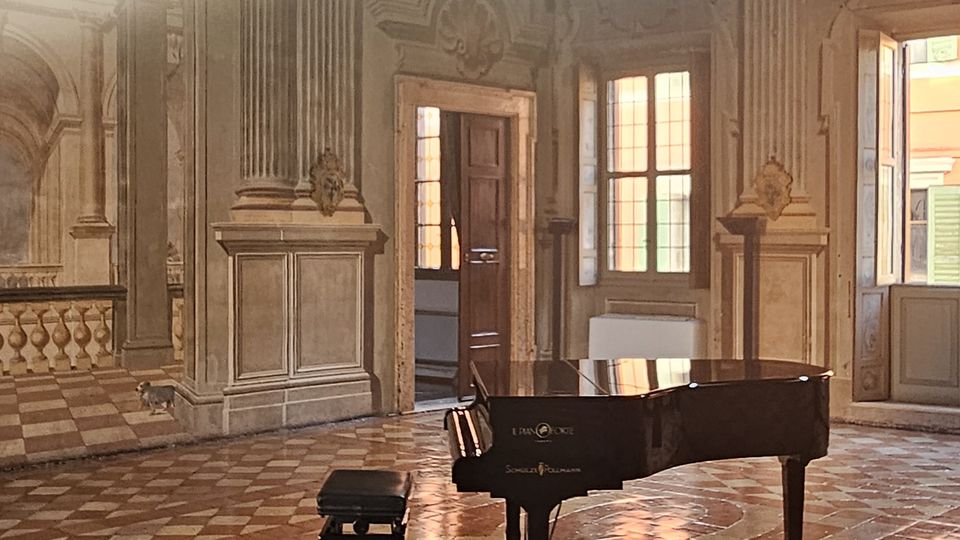 Sala storica con soffitto affrescato decorato, lampadario a gocce di cristallo appeso al centro, pavimento in piastrelle di marmo a motivi geometrici e un pianoforte a coda nero con sgabello al centro della stanza illuminata dalla luce naturale proveniente dalle finestre.