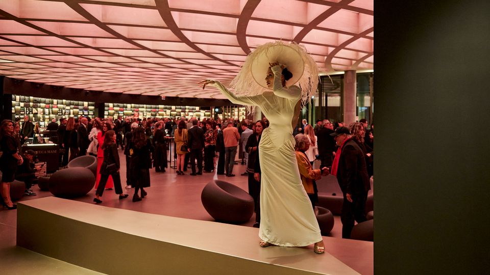 Spazio interno moderno con soffitto a griglia retroilluminato di colore rosa, arredi minimalisti e tavoli circondati da scaffali pieni di oggetti espositivi. Nell'area si trovano persone in piedi e sedute, in un contesto che suggerisce un evento o una mostra.