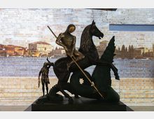 Scultura in bronzo di un cavaliere a cavallo con lancia che combatte un drago, esposta su una base in una sala interna. Sullo sfondo si vede una parete di mattoni chiari con una proiezione panoramica di un paesaggio urbano.