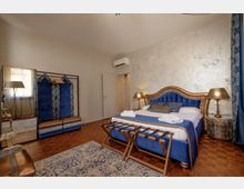 Camera da letto in stile hotel con grande letto matrimoniale con testiera imbottita blu e finiture dorate, comodini con lampade e panca portavaligie ai piedi. Pareti chiare con una parete effetto spatolato, pavimento in parquet, armadio a vista con pannelli blu e specchio, e tenda blu vicino alla finestra.