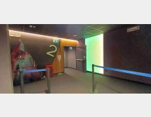 Interno di un cinema con corridoio e area d’attesa: pareti scure con grande numero “2”, segnaletica di emergenza e porta di uscita. Sono presenti transenne con nastro blu, un cestino rosso e un pannello luminoso verde su una parete laterale.