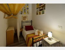 Piccola camera da letto con letto singolo in ferro battuto, copriletto a righe e comodino con lampada. A sinistra c’è un angolo guardaroba con mensola in legno e grucce, mentre sulle pareti sono presenti quadri e una nicchia con scaffali.