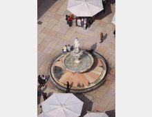 Veduta dall’alto di una piazza pavimentata in pietra con una fontana circolare al centro, con statua su piedistallo e vasca con acqua. Ai margini si notano grandi ombrelloni bianchi e una piccola area con bancarella espositiva.