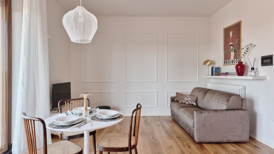 Soggiorno moderno con pavimento in legno, parete bianca con boiserie e divano grigio affiancato da una mensola con libri e decorazioni. In primo piano zona pranzo con tavolo rotondo apparecchiato, sedie in legno e lampadario a sospensione, accanto a una grande porta-finestra con tende leggere.