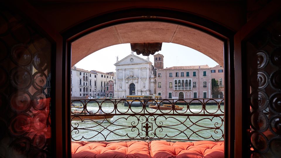Vista dal’interno di una finestra ad arco con ringhiera in ferro battuto e cuscini rossi, affacciata su un canale con imbarcazioni. Di fronte si vedono la facciata di una chiesa barocca e palazzi storici lungo l’acqua.