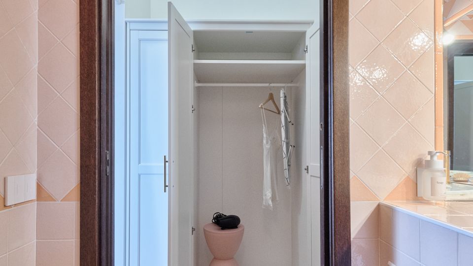 Vista attraverso una porta con cornice in legno che collega un bagno piastrellato in rosa a un piccolo spazio con pavimento in legno e un armadio bianco aperto con ripiani e gruccia. A destra si vede un piano lavabo con specchio e accessori.