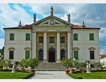 Facciata simmetrica di una villa neoclassica con frontone triangolare, quattro colonne centrali e statue sul tetto, con tetto in coppi rossi e persiane verdi. Davanti si vedono una scalinata centrale e un viale tra prato curato, vasi e piccole statue.