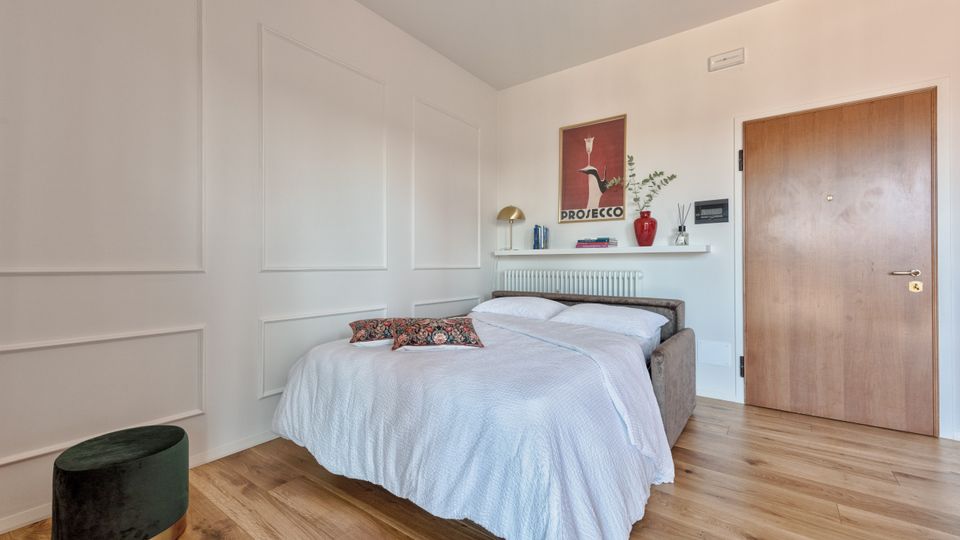Camera da letto moderna e luminosa con pavimento in legno, letto matrimoniale con copriletto bianco e pareti bianche con boiserie. Sopra il termosifone c’è una mensola con libri e decorazioni, un poster a parete e una porta in legno sulla destra.