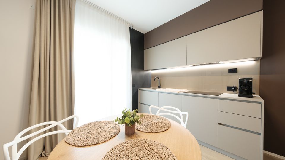 Cucina moderna e compatta con mobili bianchi a linea pulita, piano di lavoro con lavello e piano cottura illuminati da luce sotto pensile e parete marrone scuro. In primo piano un tavolo rotondo in legno con tovagliette intrecciate e sedie bianche, vicino a una grande finestra con tende chiare.