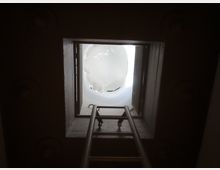Vista dal basso di un lucernario quadrato con telaio in legno, da cui entra luce intensa, con una scala metallica che conduce all’apertura. All’esterno si intravede una cupola bianca con una superficie traslucida e crepata.