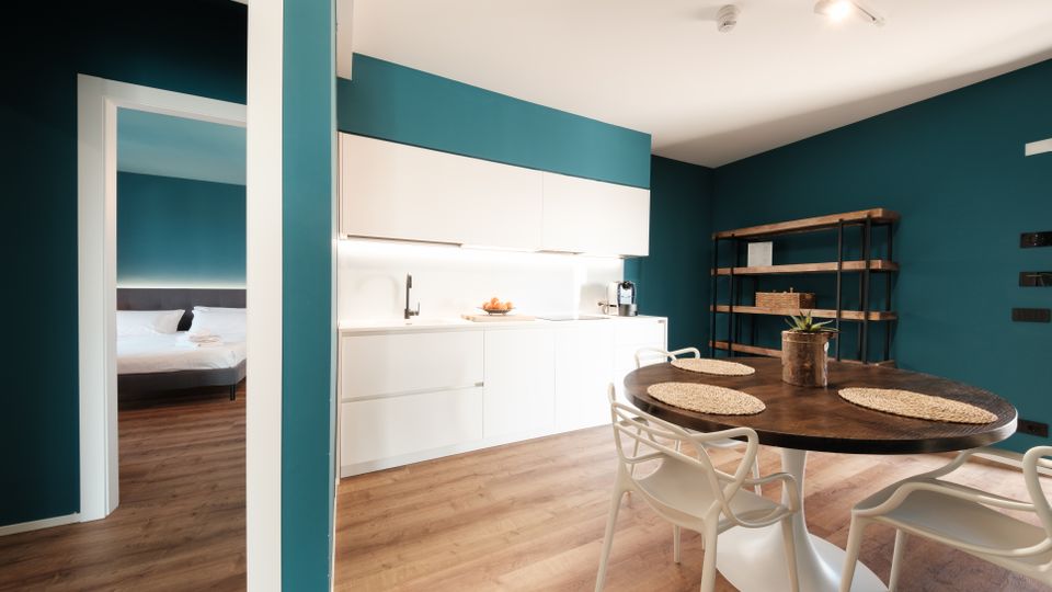 Interno di un appartamento moderno con pareti verde petrolio e pavimento in legno, con cucina lineare bianca illuminata e tavolo rotondo scuro con sedie bianche. Sullo sfondo una libreria in metallo e legno e una porta aperta che conduce a una camera da letto.