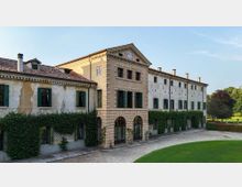 Facciata di una grande villa storica con corpo centrale in pietra e arco d’ingresso, ali laterali intonacate con finestre a scuri verdi e tetto in coppi. Davanti si vede un viale in ghiaia e un prato curato, con alberi e vegetazione sullo sfondo.