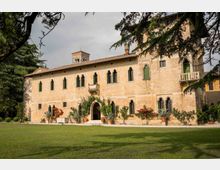 Facciata di una villa storica in pietra con finestre ad arco e persiane verdi, ingresso ad arco e piccoli balconi. Davanti si estende un prato con vasi di piante e fiori, circondato da alberi alti.