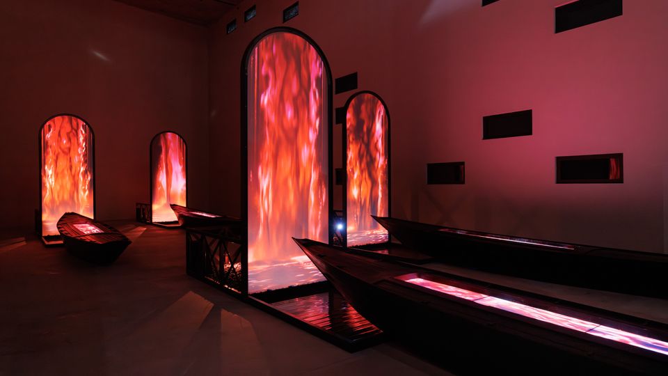 Interno di una mostra d'arte contemporanea con pareti in tonalità scura, caratterizzato da grandi installazioni verticali illuminate da luci rosse e arancioni simili a fuoco, e barche scure disposte sul pavimento.