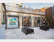 Cortile di una galleria d’arte con pavimentazione in pietra chiara e grandi porte a vetri aperte su sale espositive illuminate. All’esterno sono esposte due sculture equestri su basamenti e un dipinto appeso alla parete, con balconata in pietra al piano superiore e edifici circostanti.