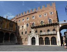 Facciata di un palazzo storico in mattoni con merlature in cima, finestre ad arco e balconi, affacciato su un’ampia piazza lastricata. Al centro si vede un portale in pietra con arco e, a sinistra, un porticato con arcate sotto un edificio adiacente.