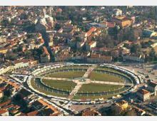 Veduta aerea di una piazza ellittica con prato centrale attraversato da viali a croce e un anello di edifici porticati che la circonda. Intorno si estende un tessuto urbano fitto con case e strade.