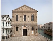 Facciata di una chiesa in mattoni a due livelli, con tre alte finestre ad arco, portale centrale incorniciato in pietra e timpano triangolare. Davanti si apre una piazza lastricata con tracciati di campo da gioco, affacciata su un canale con edifici storici lungo le rive.