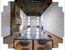 Sala riunioni lunga e stretta con grande tavolo rettangolare in vetro e sedie metalliche, pavimento in mosaico e soffitto alto a travi di legno. Sulla sinistra c’è un grande camino in pietra decorato e in fondo ampie finestre alte che illuminano le pareti bianche.