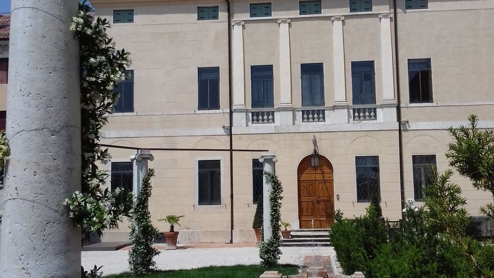 Cortile con prato e vialetto in mattoni che conduce a una fontana bassa davanti a un edificio in stile classico con frontone, alte finestre e portone centrale ad arco. In primo piano si vede una colonna in pietra e siepi curate ai lati del percorso.