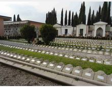 Cimitero con file di lapidi basse in pietra e vialetti di ghiaia, disposto su prati a gradoni. Sullo sfondo si vedono cappelle funerarie bianche con croci e un lungo filare di cipressi.