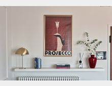 Parete interna di un ambiente domestico con mensola bianca sopra un radiatore, decorata con lampada da tavolo in ottone, libri, diffusore e vaso rosso con rami verdi. Sopra la mensola è appeso un grande poster incorniciato con scritta “PROSECCO” su fondo rosso.