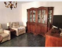 Soggiorno con due poltrone beige e un tavolino con lampada, pavimento scuro e pareti chiare. Sul fondo c’è una grande vetrina in legno con ante in vetro e, a destra, un mobile in legno con televisore; dal soffitto pende un lampadario classico.