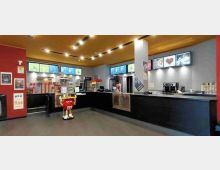 Interno di un chiosco bar/ristoro con bancone a L nero, vetrine espositive, macchine del caffè e dispenser di bevande. Sulle pareti ci sono menu digitali illuminati e il soffitto in legno con faretti, in un ambiente moderno con pavimento grigio.