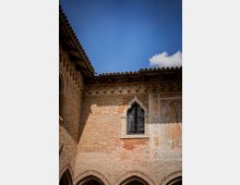 Cortile di un edificio storico in mattoni con archetti decorativi sotto la gronda e una finestra gotica con inferriata. A destra si vede un affresco parziale sulla parete, sotto un cielo azzurro.