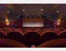 Interno di una piccola sala cinematografica con file di poltrone imbottite e uno schermo centrale incorniciato da tende rosse. Pareti rivestite e soffitto basso con luci incassate creano un ambiente raccolto e simmetrico.