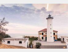 Faro costiero bianco con lanterna e balconata circolare, affiancato da un edificio basso con tetto in tegole rosse e persiane scure. L’insieme si trova su un’area recintata vicino al mare, con dune sabbiose e cielo nuvoloso sullo sfondo.