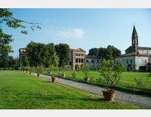 Ampio giardino con prato curato e vialetto in ghiaia fiancheggiato da grandi vasi di terracotta con piante. Sullo sfondo si vede un complesso di edifici storici in pietra e intonaco, con un campanile a guglia sulla destra e alberi che incorniciano le facciate.