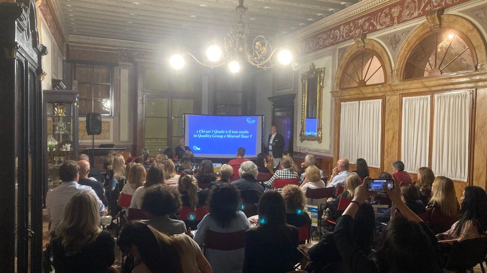 Sala conferenze in un palazzo storico con soffitto a travi dipinte, lampadario centrale e pareti decorate con boiserie e archi con tende bianche. In fondo alla stanza è visibile un grande schermo per presentazioni affiancato da un leggio, con file di sedie rivolte verso lo schermo.