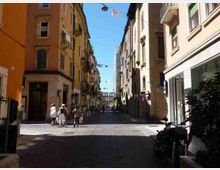Strada cittadina stretta con pavimentazione in pietra, fiancheggiata da alti edifici residenziali dai colori caldi con balconi e vetrine al piano terra. Lampade sospese attraversano la via e, in fondo, si intravede un passaggio o ponte tra gli edifici sotto un cielo sereno.