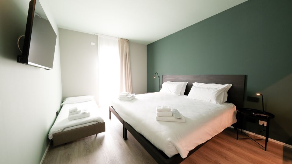 Camera da letto moderna con letto matrimoniale e letto singolo, biancheria bianca e asciugamani piegati. Parete verde scuro con testiera in legno, TV a parete, comodini con lampade e grande finestra con tende che illumina un pavimento in legno chiaro.
