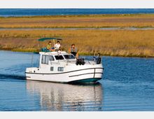 Barca cabinata bianca che naviga in un canale d’acqua calma, con ponti e finestrature e una piccola copertura superiore. Sullo sfondo si estende una zona umida con canneti e ampie distese d’acqua.