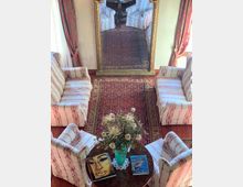 Salotto con pavimento in legno e due tappeti persiani, arredato con quattro poltrone e due divani a righe disposti intorno a un tavolino rotondo con vaso di fiori e libri. Sul fondo c’è un grande specchio con cornice dorata tra due finestre con tende rosse.