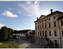 Facciata di una villa storica in stile neoclassico con frontone, grandi finestre e scala esterna in pietra con ringhiere, affacciata su un cortile in ghiaia. A lato si vede un edificio basso con portici ad archi e vegetazione, sotto un cielo azzurro con nuvole sparse.