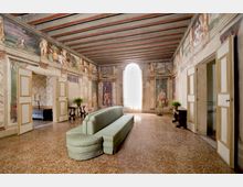 Ampio salone interno in stile storico con pareti affrescate e soffitto a travi, pavimento in terrazzo e grande finestra ad arco sul fondo. Al centro c’è un divano lungo verde chiaro, con porte laterali aperte e piccoli tavoli con piante lungo le pareti.