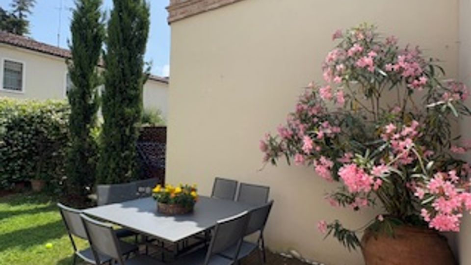 Piccolo giardino con tavolo da esterno e sedie in metallo su un patio, accanto a un muro color crema con bordo merlato. Sono presenti cipressi, prato verde e un grande vaso con arbusto fiorito rosa.
