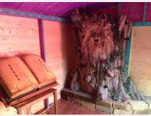 Interno di una piccola struttura in legno con pareti colorate e soffitto viola, con un leggio a sinistra. In fondo c’è una grande scultura decorativa simile a un albero con volto, circondata da fogliame e pietre su una base rialzata.